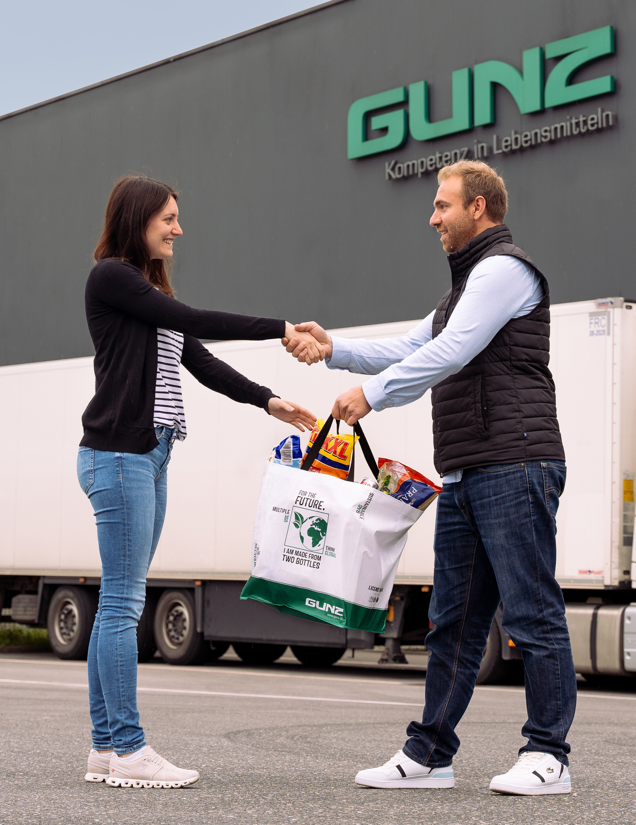 Twee personen die elkaar de hand schudden voor het hoofdkantoor van Gunz, een boodschappentas met producten wordt overhandigd, een vrachtwagen op de achtergrond