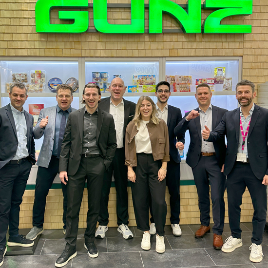 ISM-Team vor GUNZ-Logo, l&auml;chelnd, in Business-Outfits vor S&uuml;&szlig;warenregal